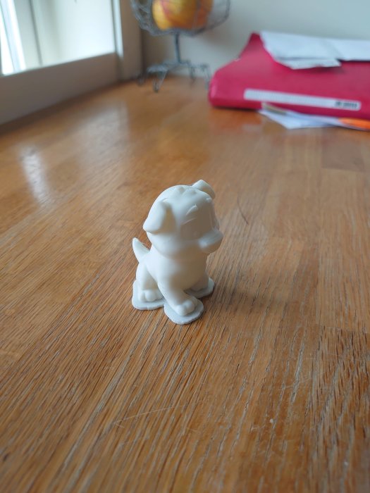 3D-utskriven vit liten hundfigur på trägolv framför fönster och dokument.