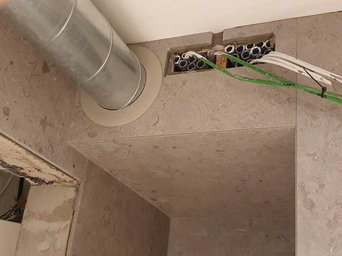 Ventilationsrör med täckbricka installerad i taket intill kakel, med synliga kablar och rörledningar vid sidan.