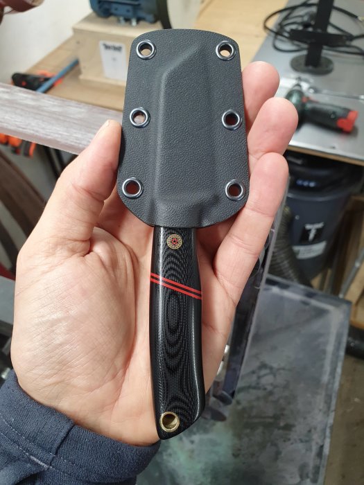 Hand håller en nygjord kniv med svart handtag och rött streck, samt ett svart knivfodral.