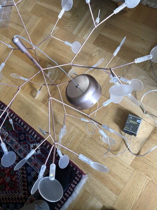Modern lampa med flera armar och ljuskällor som står på ett trägolv, sladd hänger löst kopplad till en transformator.