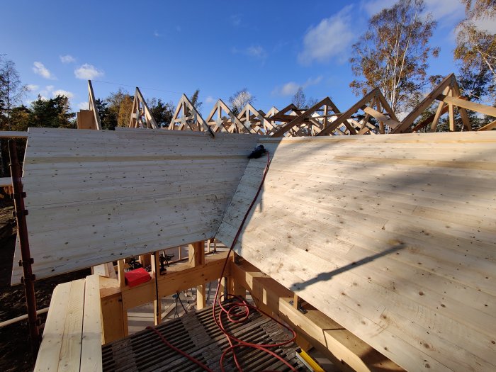 Byggplats med halvfärdigt trähus under konstruktion, synliga takstolar och pågående råspontsarbete under klar himmel.