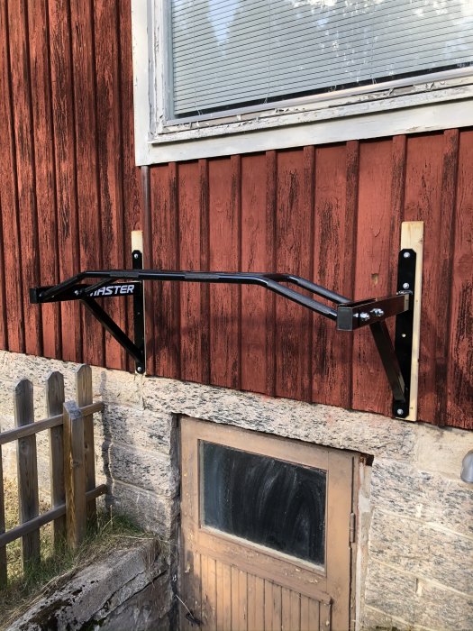 Svart pull-up bar monterad på en röd trävägg utomhus bredvid ett fönster.