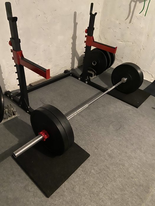 Hemmagym med skivstång och vikter på golvmattor, squat rack i bakgrunden mot en vit vägg.