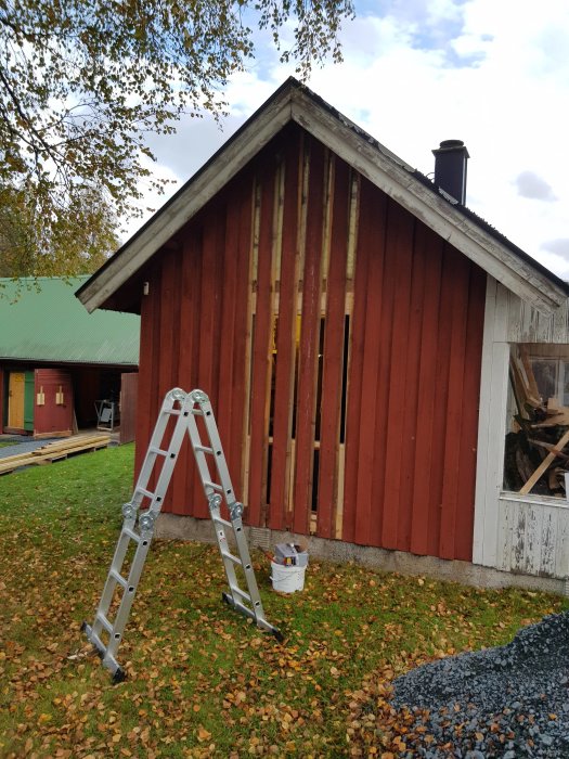 Gäststuga med nymonterat fönster, röd och vit panel under renovering, stege och hink med falufärg framför byggnaden.