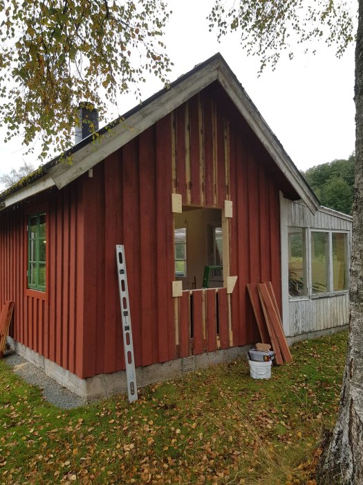 Röd stuga under renovering med nyinstallerat fönster, träpanel och fönsterbleck, stegar och verktyg synliga.