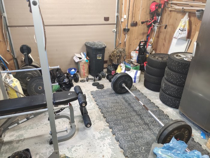 Hemmagym med träningsbänk och vikter på gummimatta i ett stökigt garage.