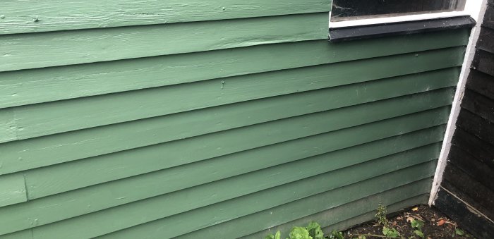 Sida av grönt garage med plankor utan synlig grusram, nära jord och vegetation.