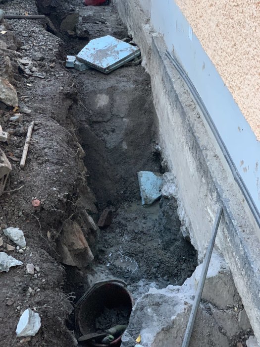 Grävd grop vid husgrund med stenar och vattenansamling, övervägande pumpbrunn för dränering.