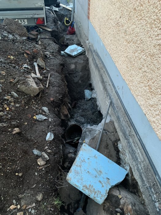 Grävd schaktning längs husgrund med synligt vattenansamling och stora stenar, spade i förgrunden.