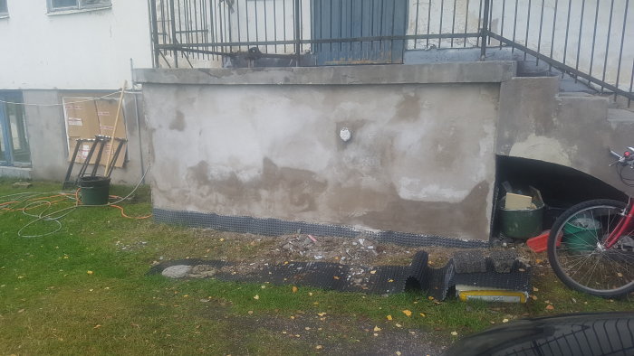 Ojämnt putsat grundmur på hus med verktyg och byggmaterial spridda på gräset.