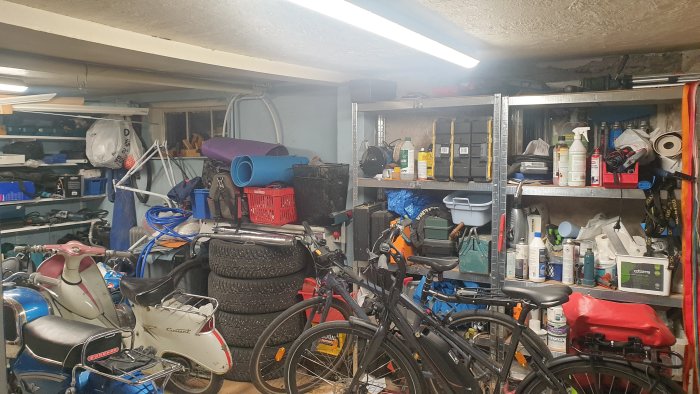 Rörigt garage med cyklar, däck och hyllor fulla av föremål, synliga takbalkar som böjer sig.