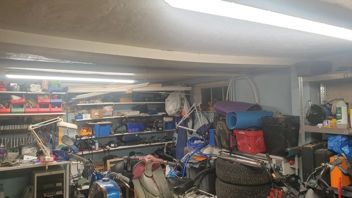 Rörigt garage med bristfälligt tak där puts har lossnat, lagringshyllor och diverse föremål.