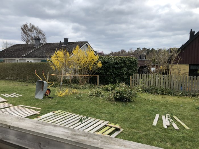 Nybyggt armeringsnätstaket längs trädgård med skottkärra, träplankor och blommande buskar i förgrunden.