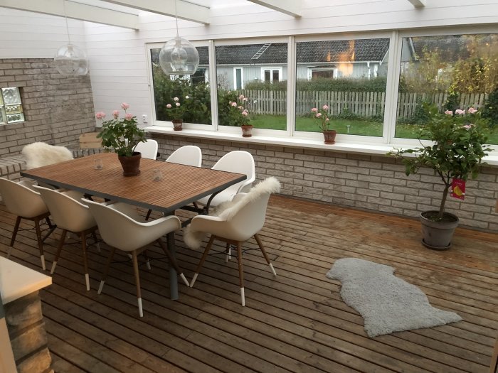 Inglasad veranda med ett trädäck, matbord och stolar, omgiven av vitmålade tegelväggar och fönster med utsikt över trädgård.