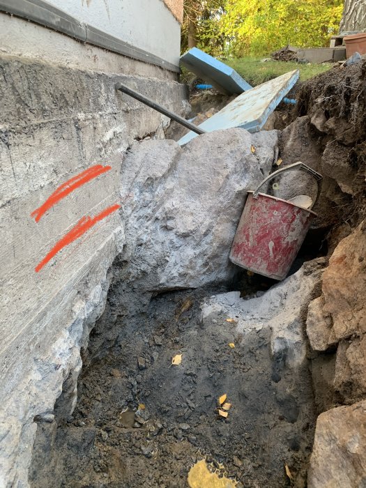 Grävd grop vid husgrund med synlig betongplatta och röda markeringar, omgiven av stora stenar och byggmaterial.