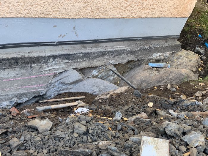 Grävd grund vid husvägg med synlig golvplatta och rosa märklinjer i en byggmiljö.