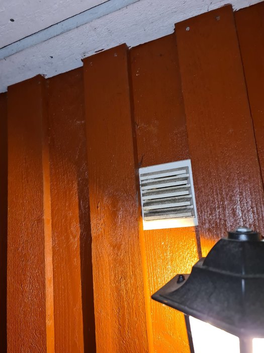 Tilluftsventil på en orangefärgad vägg i ett hörn nära taket med en lampa i förgrunden.