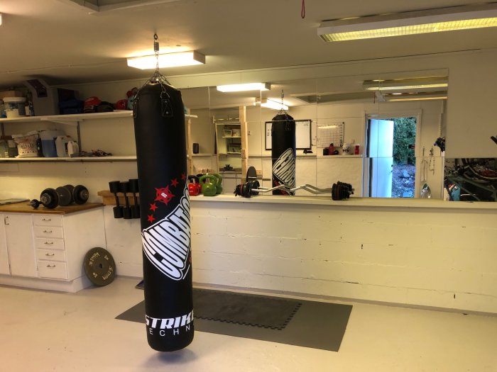 Hemmagym med svart sandsäck, hantlar på hylla, gröna kettlebells och viktplatta på golv med epoxi.