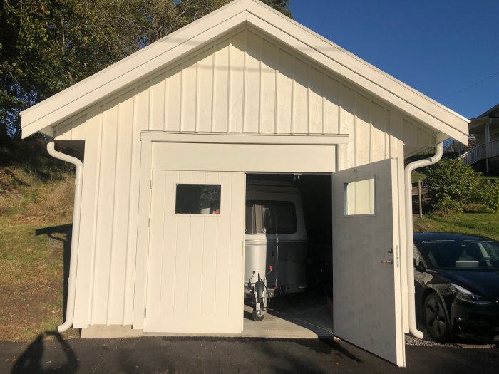 Vit garagebyggnad med öppen tung garageport som avslöjar en husvagn inuti, byggd med reglar och plywood.