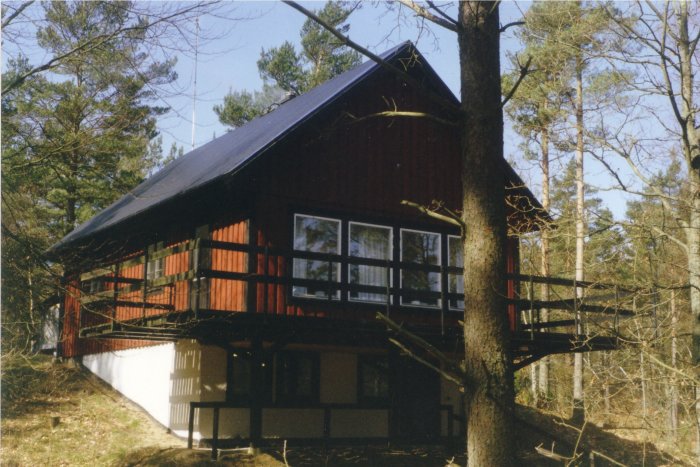 Trähus med suterrängkällare byggt i skogsmiljö, med veranda och stora fönster.