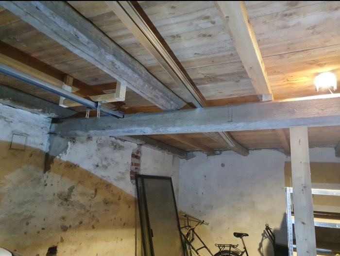 Gammelt bjælkelag i et loft med træbjælker og murværk, understøttet af en ny stolpe under midterbjælken; baggrunden viser cykler og reoler.