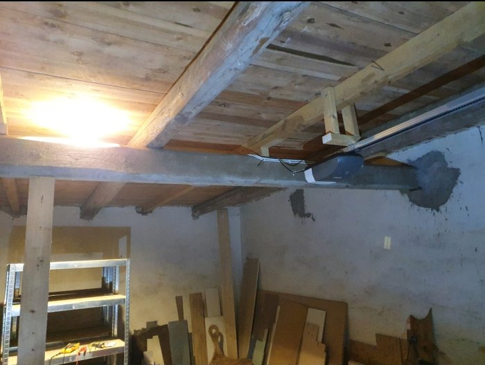 Loft med træbjælker og en stolpe, der støtter en tværgående bjælke. Rustik mur med hylder og værktøj i baggrunden. Loftet skal omdannes til træningsrum.