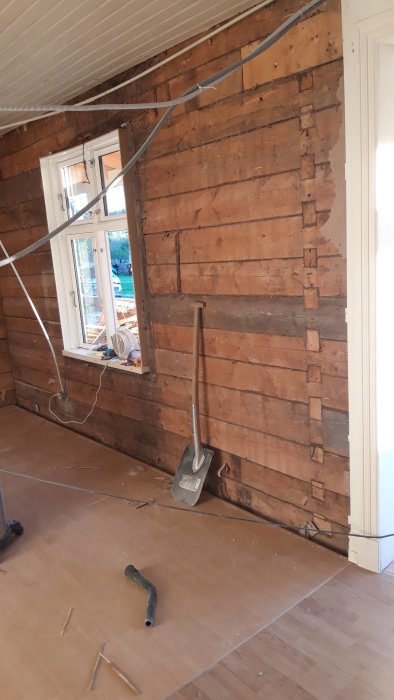 Interiörbild av en renoveringsprocess med synliga liggande träplank på en vägg, omgivet av verktyg och renoveringsmaterial.