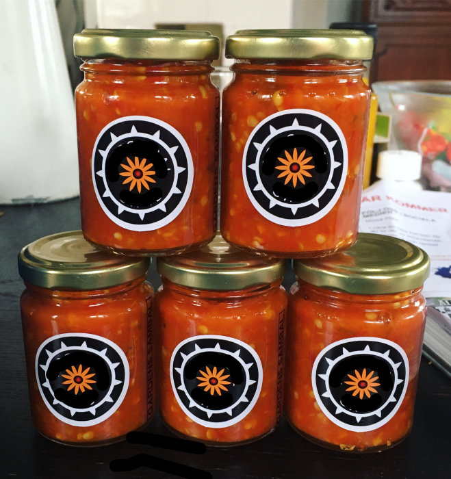 Sex små glasburkar med hemlagad sambal, märkta med etiketter som har en blomliknande symbol.