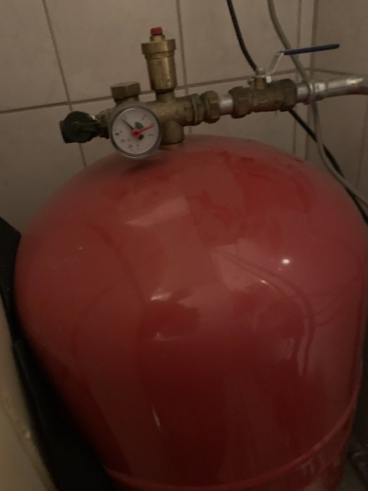 Manometer på toppen av en röd ackumulatortank för värmesystem i ett dunkelt utrymme.