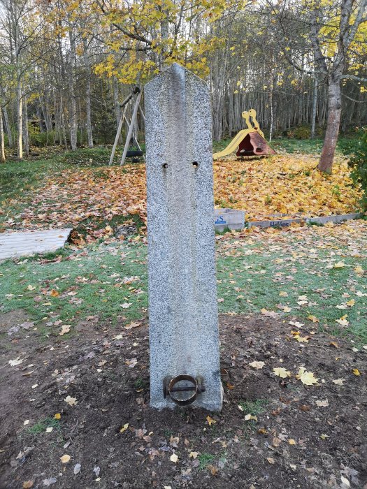 En flaggstångssten i granit står på en gräsmatta med höstlöv och en lekställning i bakgrunden.