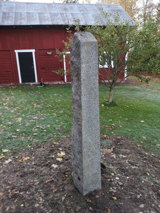 Hög flaggstångssten av granit nergrävd i trädgård framför röd byggnad, omgiven av gräs och höstlöv.