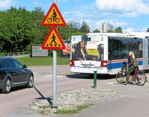 Cykelvägskylt och gångvägskylt vid korsning med buss, bil och cyklist i rörelse.