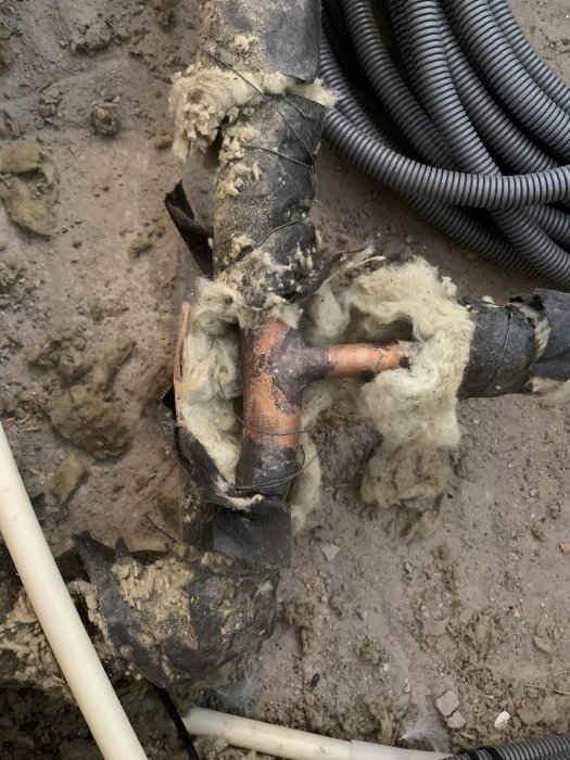 Äldre kopparledning i ett byggprojekt isolerad med glasull, omgiven av grus och elektriska ledningar.