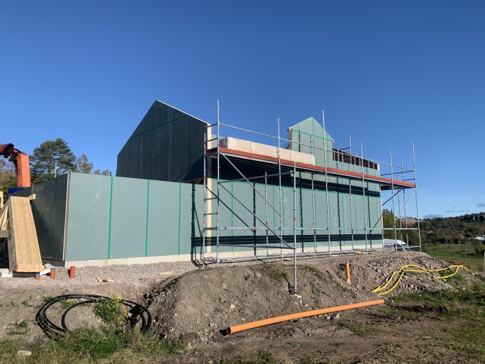 Hus under konstruktion med takstolar och övervåning omgiven av byggställningar mot blå himmel.