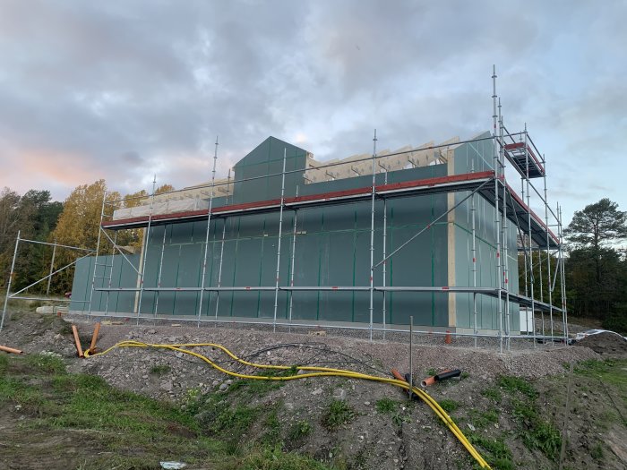 Nybyggt hus med ställningar, otäckt tak och skog i bakgrunden.