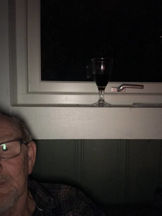 En person halvt synlig i bild med ett glas vin på fönsterbrädan framför ett öppet fönster i mörkret.