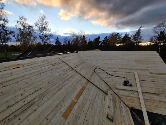 Halvfärdigt träsponttak med verktyg och byggmaterial i skymningen, med himmel och träd i bakgrunden.