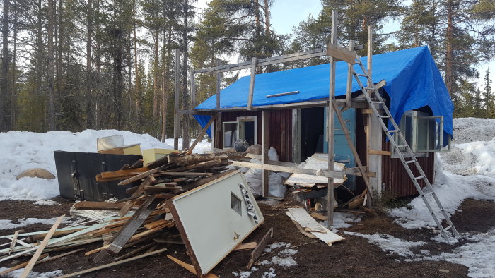 Rivningsarbete av våningssängar, med högar av byggavfall framför stuga under blå presenning.