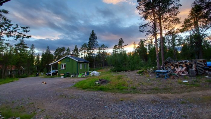 Hus i skogsglänta vid solnedgång, ojämn gräsmatta, ny trappa av återanvänt trallvirke, vedstapel.