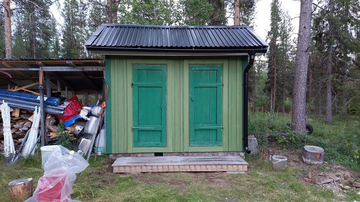 Trädgårdsbod med gröna dörrar och ny trapp av återanvänt trallvirke framför, omges av skog.