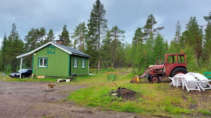 Grönmålat hus med veranda, en hund i förgrunden, röd traktor och vita plaststolar utomhus.