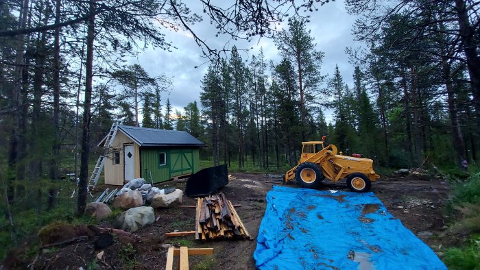 Nybyggt skogsstuga med grön fasad, vit dörr och stege mot huset, bredvid en gul hjullastare.