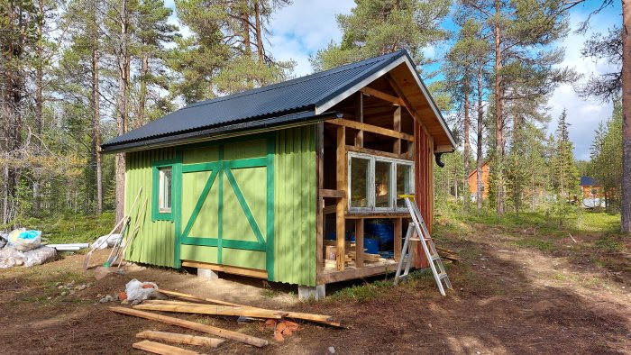 Halvfärdigt byggprojekt av trähus med pågående arbete, grönmålad vägg och gammal dörr, i skogsområde.