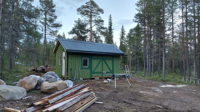 Nyinstallerad grön dörr på ett nyligen renoverat skjul med skogsbakgrund och byggmaterial i förgrunden.