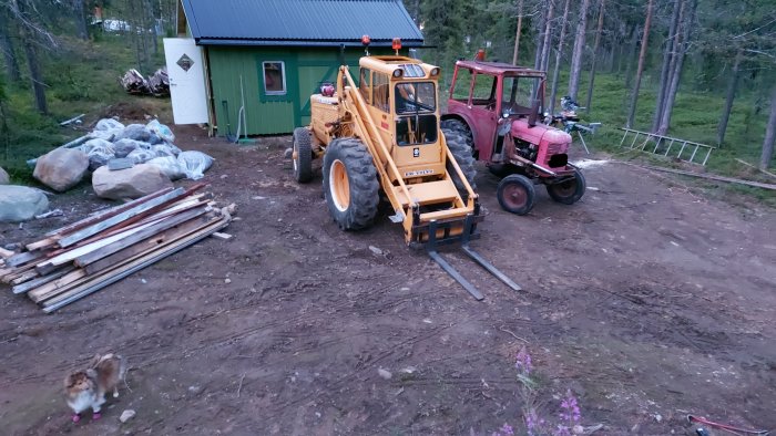 Byggarbetsplats med en gul hjullastare, en röd traktor och en hund, framför ett grönt hus med byggmaterial.