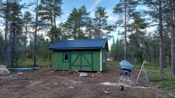 Nybyggt grönt skjul med svart tak i skogsmiljö, byggverktyg syns i förgrunden.