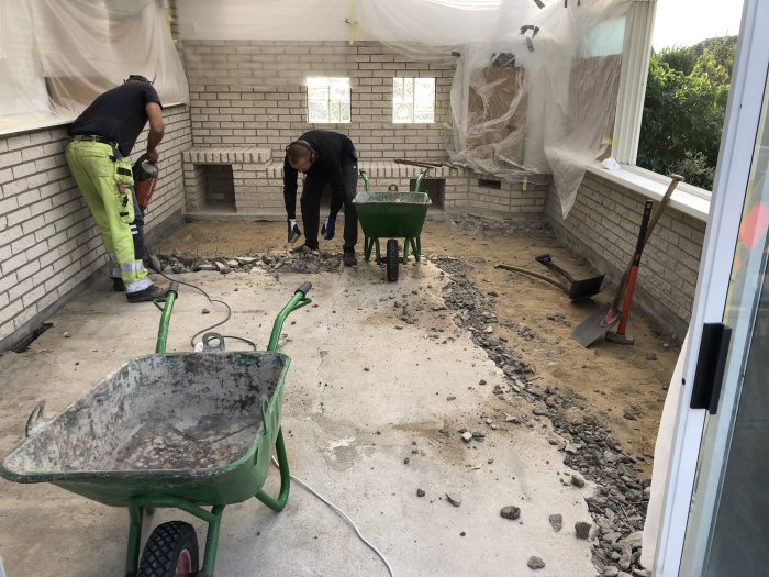 Renovering av uterum med arbetare som gräver upp betonggolv och lera, omgivna av byggverktyg och skottkärror.