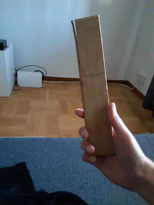 Hand som håller upp en skadad parkettbit framför ett laminatgolv av liknande mönster i ett vardagsrum.