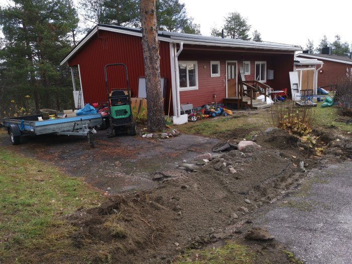 Röd stuga med utspridd 0-18 stenmaterial vid grävd fiberdike nära hus och släpvagn.