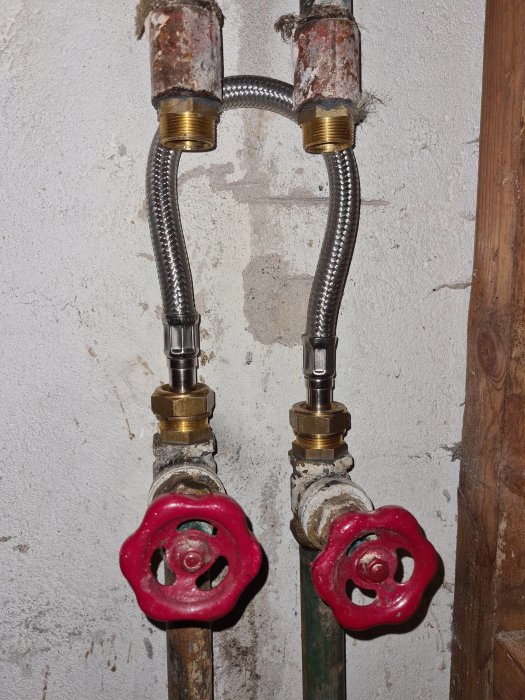 Två kopparledningar med flexibla anslutningar och röda ventiler på en betongvägg i ett garage.